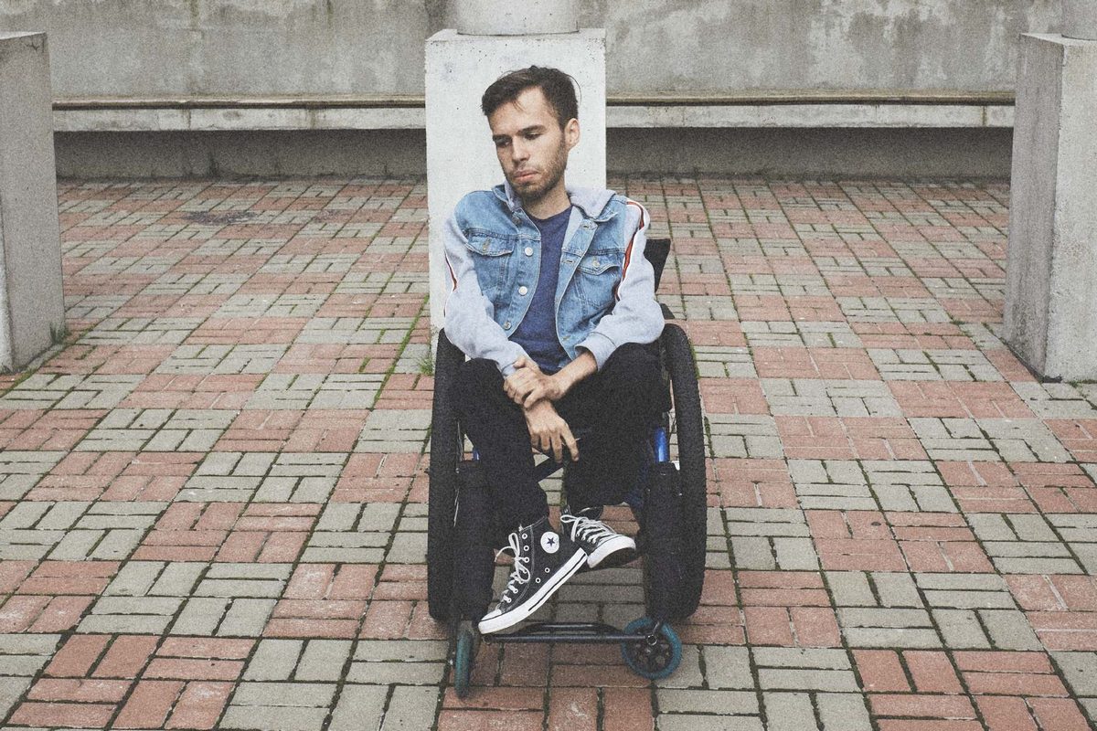  На фото молодой мужчина в инвалидной коляске на фоне городского пейзажа.