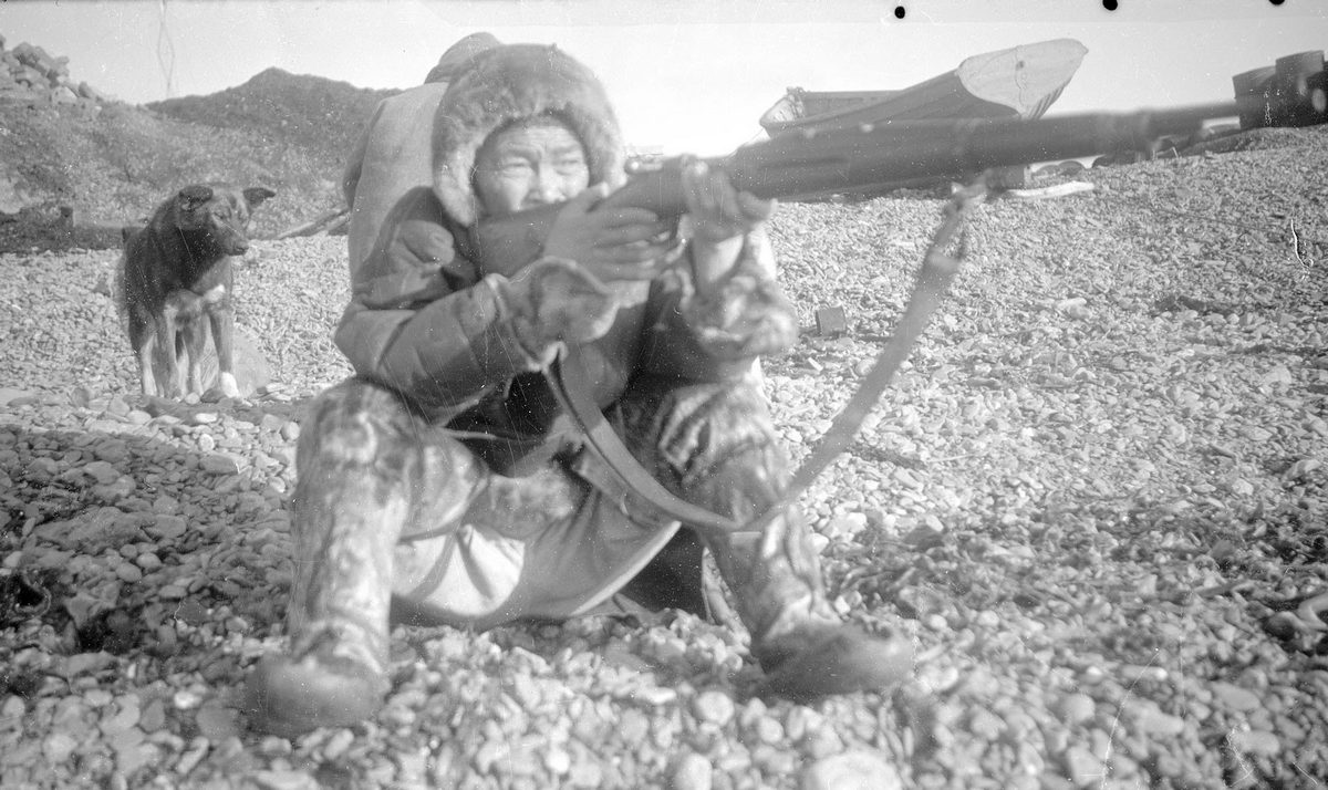  На старой черно-белой фотографии мужчина-ненец целится куда-то из винтовки, сидя на галечном пляже. Позади него собака и лодки.